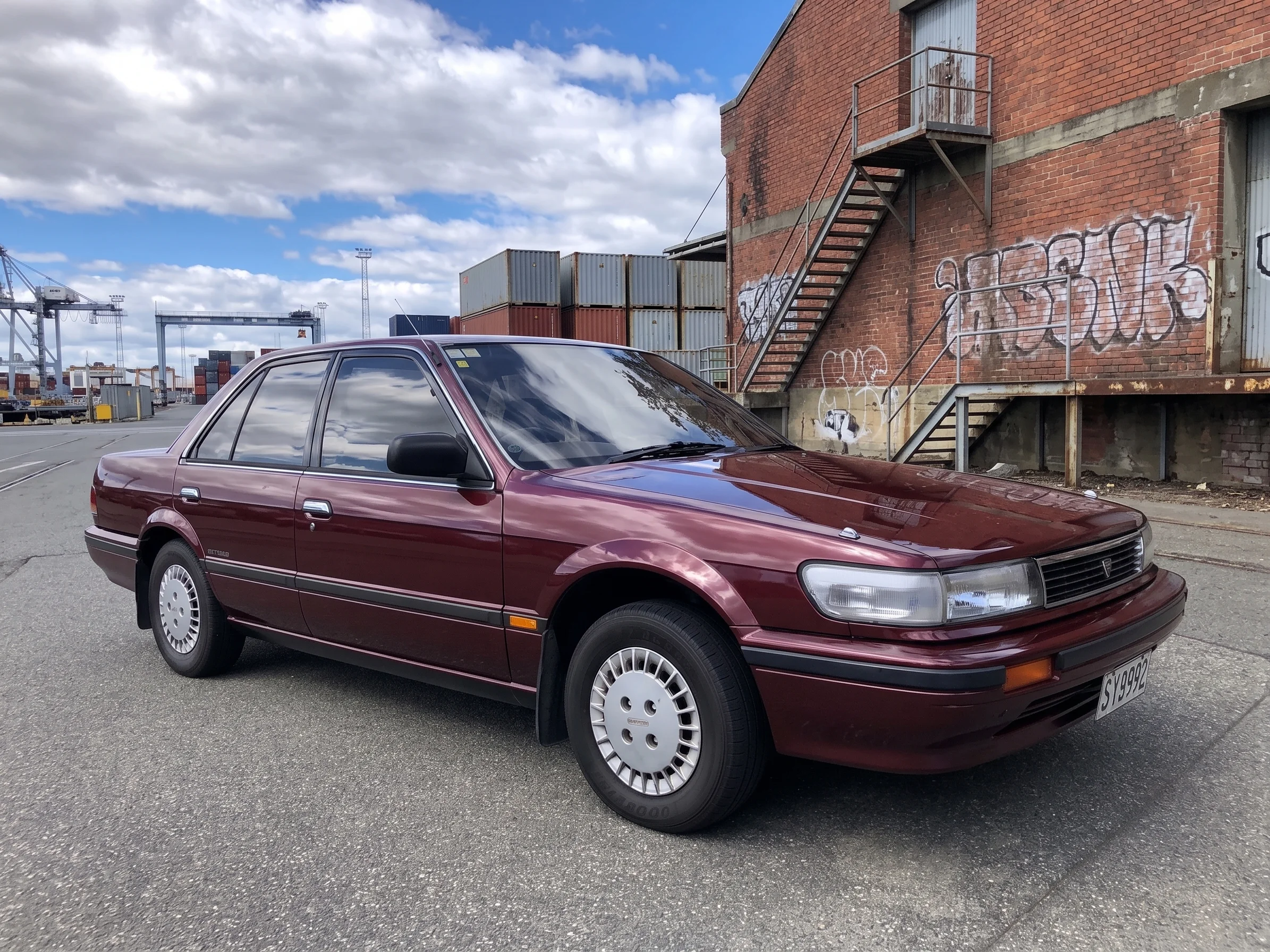 A vintage dark red sedan parked on an asphalt road, featuring an urban industrial background with a red brick building, metal stairs, and shipping containers under a cloudy sky.