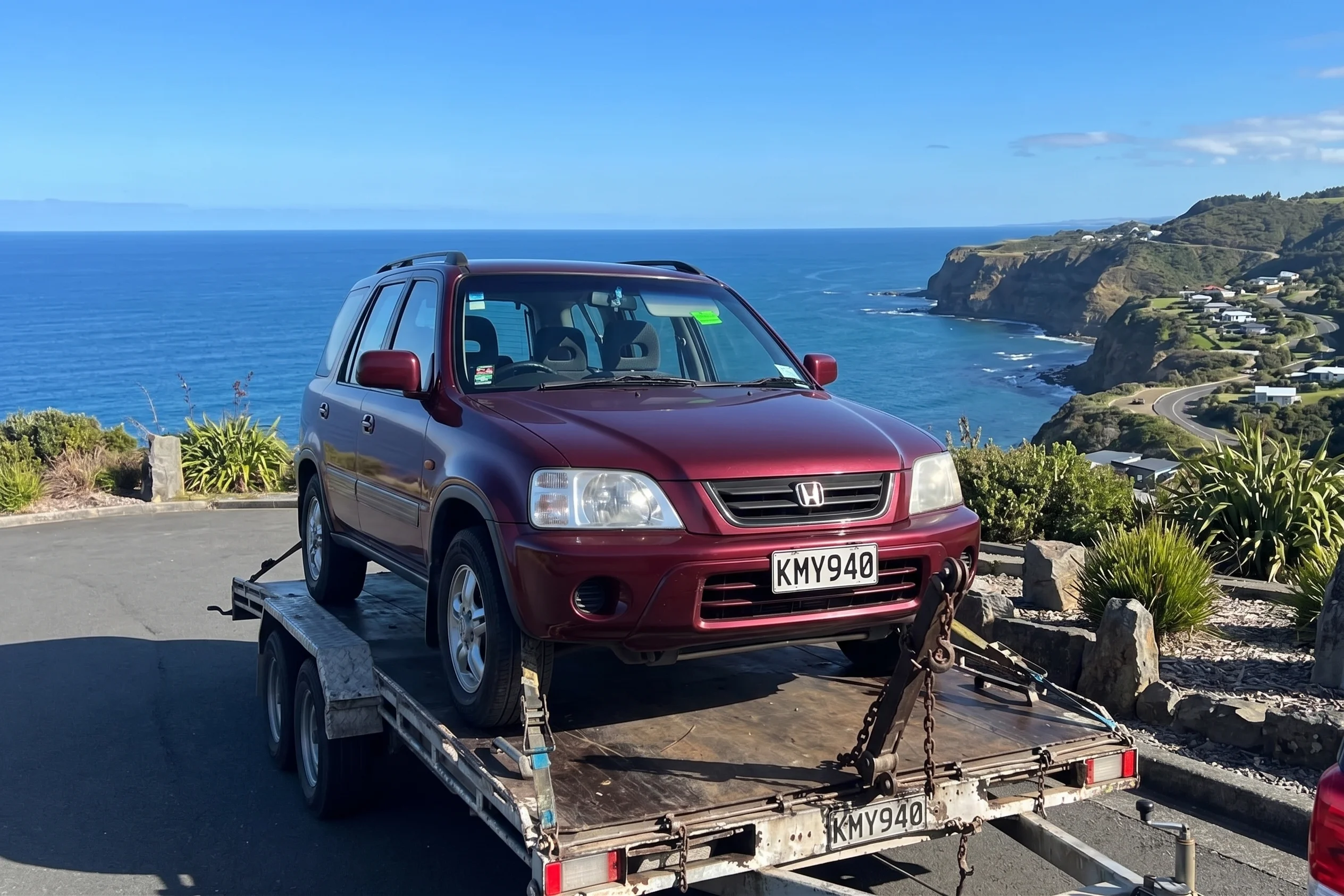 A maroon Honda CR-V SUV loaded on a trailer, parked at a viewpoint overlooking a cliffside and the sea.